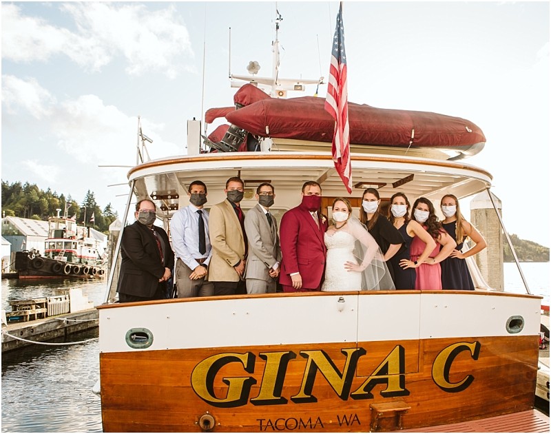 A group of people wearing masks pose together on the deck of the boat gina c docked at a snohomish marina with an american flag visible and water and trees in the background Snohomish wedding photography