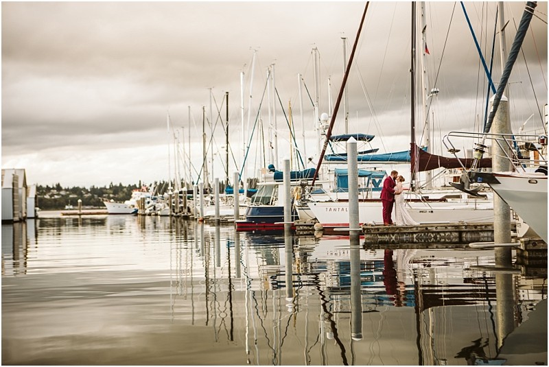 A snohomish marina scene with several docked sailboats on calm water Two people in red stand on a dock beside a white sailboat with reflections of boats and masts shimmering under a cloudy sky Snohomish wedding photography