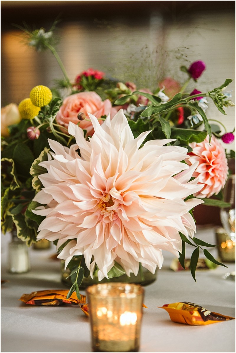 A close up of a table centerpiece at a snohomish event featuring a large pale pink dahlia greenery and colorful flowers In the foreground a lit votive candle and wrapped candies rest on a white tablecloth Snohomish wedding photography