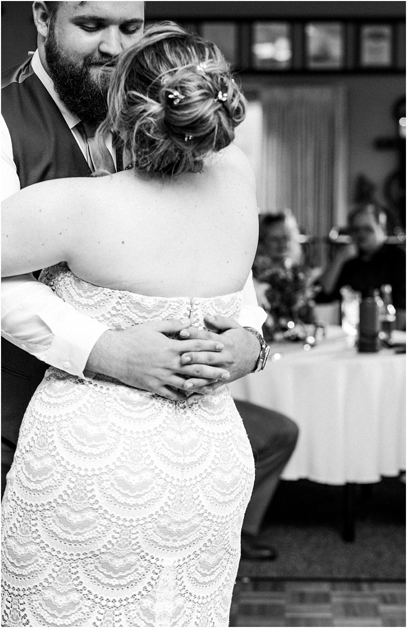 A bride and groom share a close embrace while dancing at their snohomish wedding reception The bride wears a lace strapless dress and the groom has a beard and vest Guests are seated at tables in the background Black and white photo Snohomish wedding photography