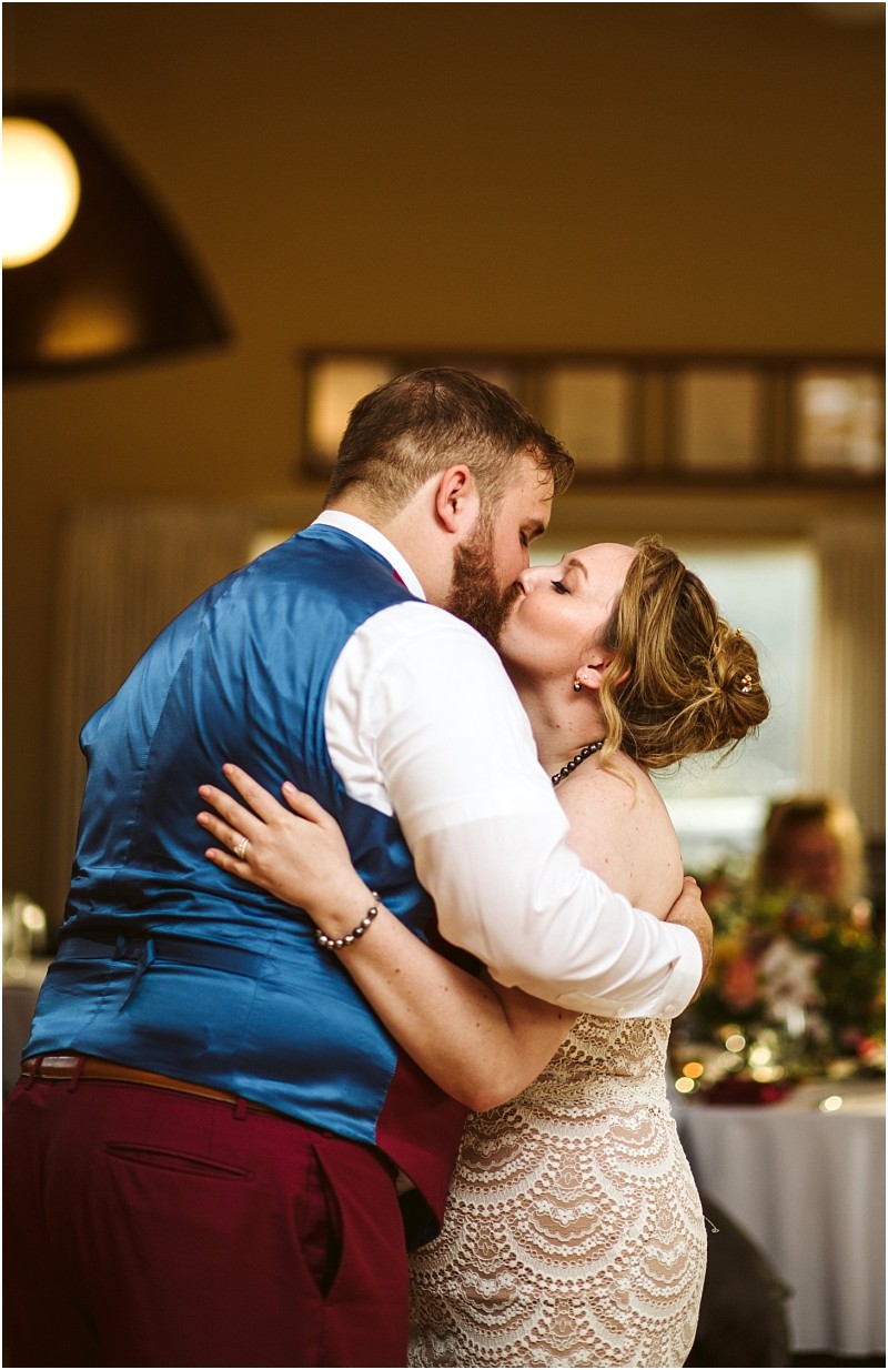 A bride and groom share a kiss during their first dance at a snohomish wedding reception embracing closely in a warmly lit room The bride wears a lace dress and the groom wears a blue vest and maroon pants Snohomish wedding photography