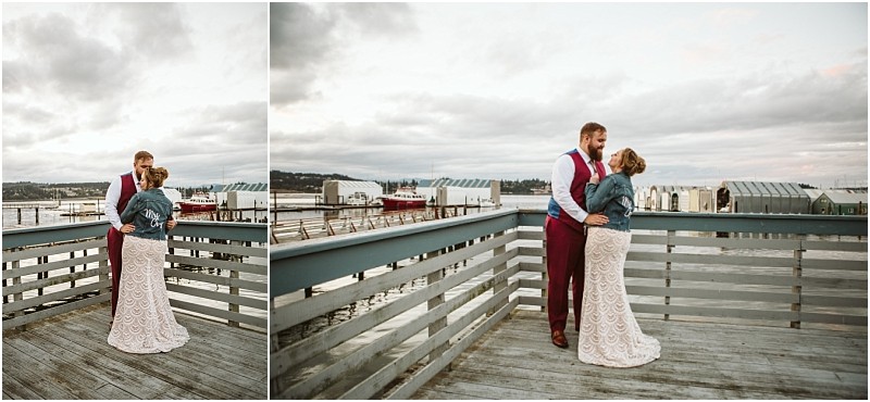 A couple stands on a wooden dock by the water under a cloudy sky in snohomish The bride wears a white lace dress and denim jacket while the groom is in a burgundy suit They embrace with boats and buildings in the background Snohomish wedding photography