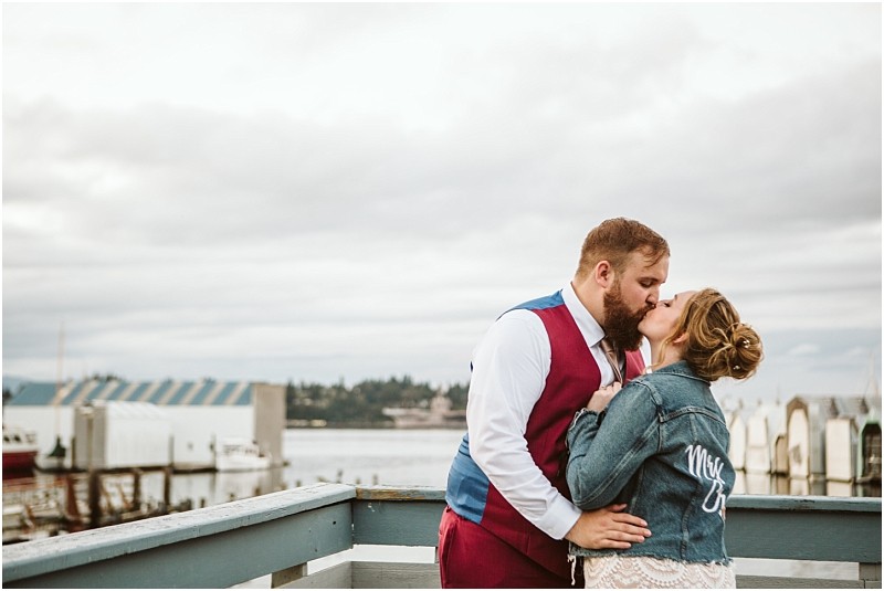 A couple kisses on a snohomish waterfront balcony the woman wears a denim jacket with mrs on the back while the man sports a suit with a red vest The background reveals water docks and cloudy skies Snohomish wedding photography