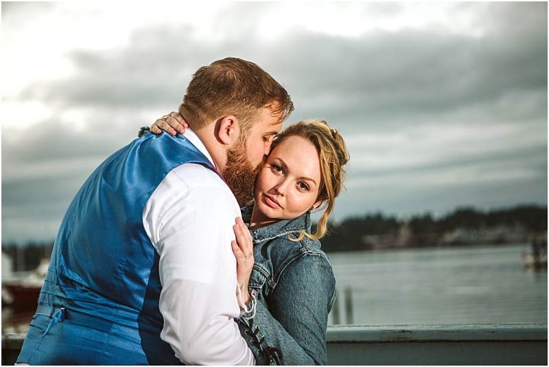 A man in a blue vest kisses a woman in a denim jacket on the cheek as she looks at the camera They stand close together outdoors by the water in snohomish with a cloudy sky and shoreline in the background Snohomish wedding photography