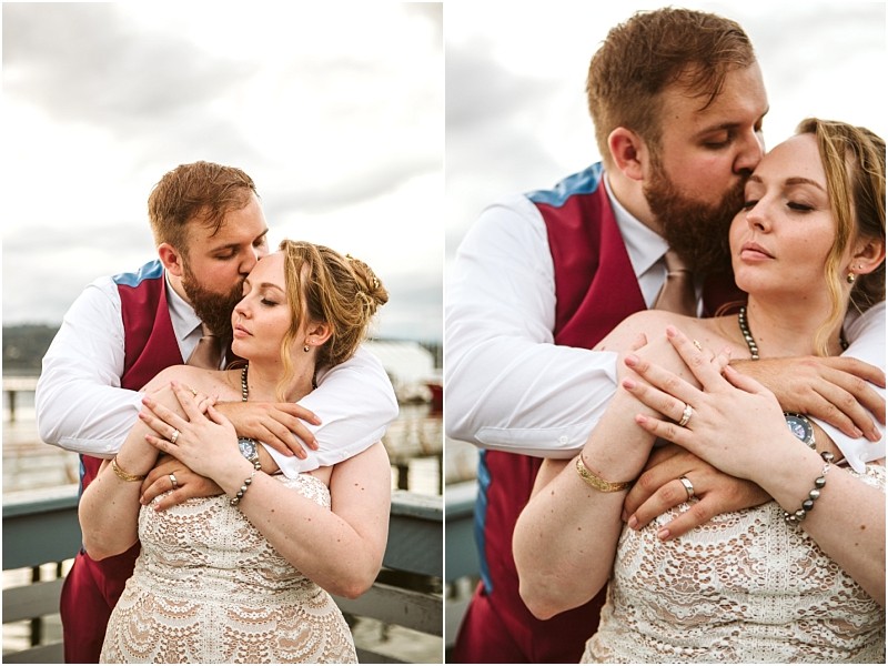 A groom in a burgundy vest embraces and kisses his bride who wears a lace dress They share an intimate moment by a railing under cloudy skies capturing the romance of their snohomish wedding Snohomish wedding photography