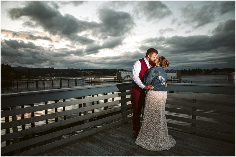 A couple stands on a wooden deck by the water at sunset in snohomish The woman wears a lace dress and a denim jacket reading mrs  as they embrace and kiss under a dramatic cloudy sky Snohomish wedding photography