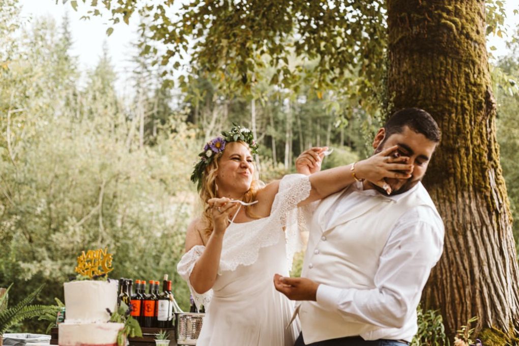 A bride playfully smears cake on the grooms face during a joyful outdoor snohomish wedding Both are smiling as they stand by a cake and bottles surrounded by lush trees and greenery Snohomish wedding photography A bride playfully smears cake on the grooms face during a joyful outdoor snohomish wedding Both are smiling as they stand by a cake and bottles surrounded by lush trees and greenery Snohomish wedding photography