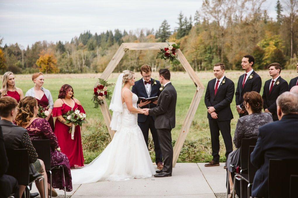 A bride and groom stand under a wooden hexagonal arch holding hands during an outdoor snohomish wedding ceremony surrounded by their wedding party and guests with trees and fields in the background Snohomish wedding photography A bride and groom stand under a wooden hexagonal arch holding hands during an outdoor snohomish wedding ceremony surrounded by their wedding party and guests with trees and fields in the background Snohomish wedding photography