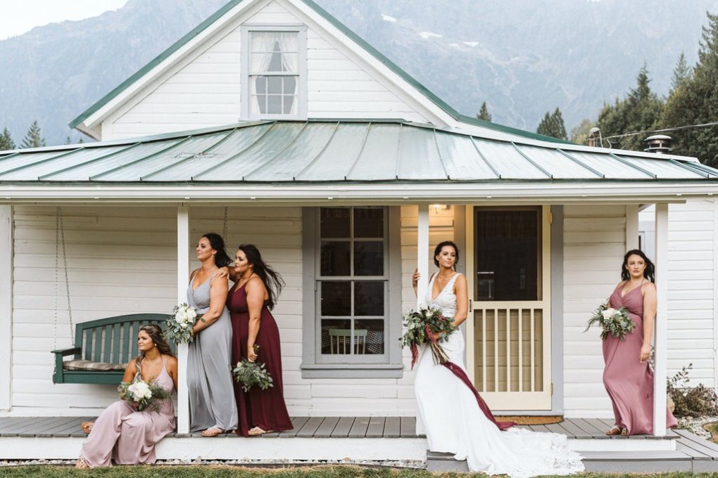 Five women in dresses pose with bouquets on the porch of a white house with a green roof in snohomish set against a backdrop of mountains and trees One woman wears white while the others wear mismatched gowns in muted tones Snohomish wedding photography Five women in dresses pose with bouquets on the porch of a white house with a green roof in snohomish set against a backdrop of mountains and trees One woman wears white while the others wear mismatched gowns in muted tones Snohomish wedding photography