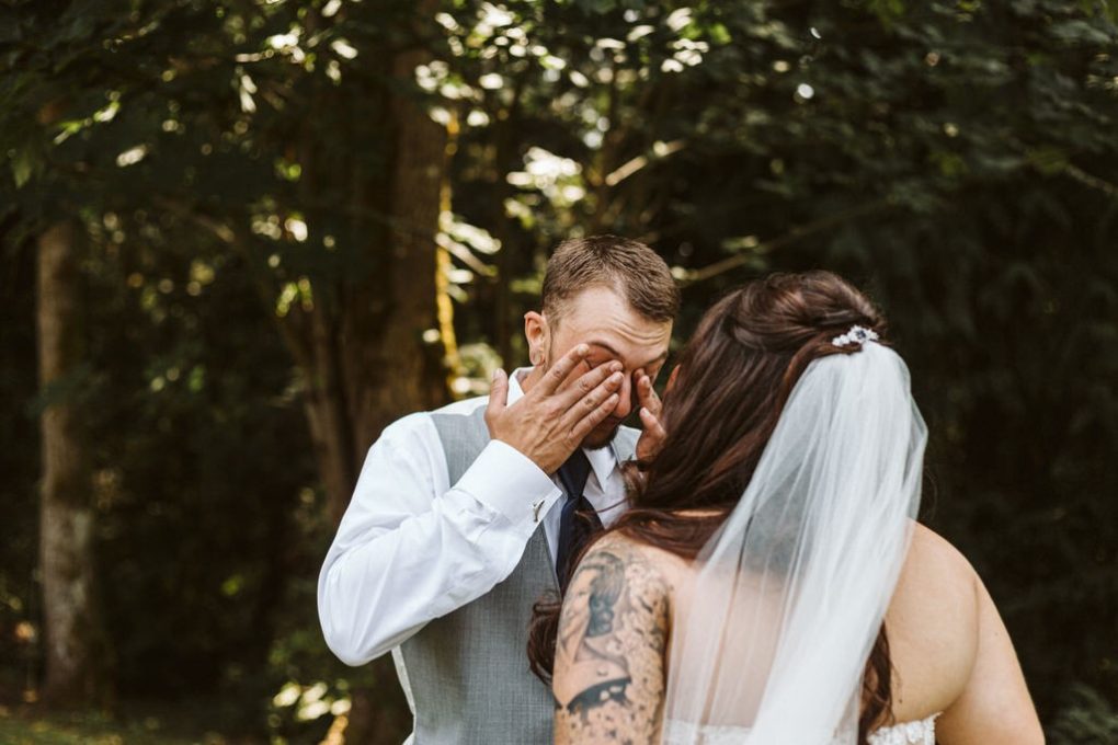 A groom wipes tears from his eyes while standing outdoors with a bride in a white dress and veil The brides back showing a tattoo and long dark hair faces the camera as sunlight filters through trees in scenic snohomish Snohomish wedding photography A groom wipes tears from his eyes while standing outdoors with a bride in a white dress and veil The brides back showing a tattoo and long dark hair faces the camera as sunlight filters through trees in scenic snohomish Snohomish wedding photography