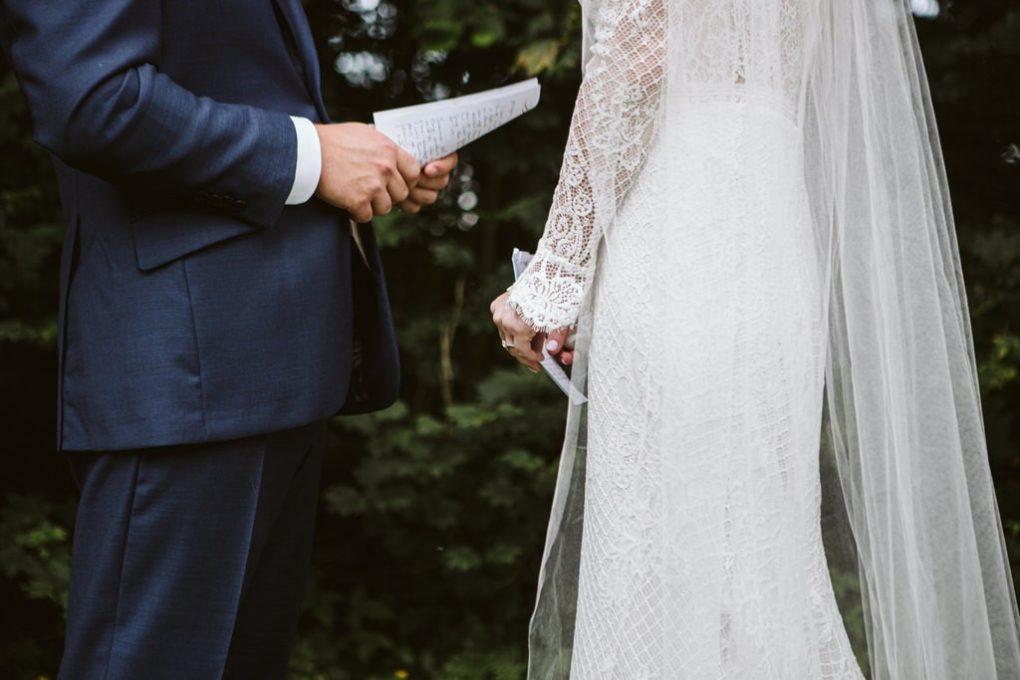 A bride in a lace wedding dress and veil stands next to a groom in a navy suit Both are holding sheets of paper possibly wedding vows with lush snohomish greenery blurred in the background Snohomish wedding photography A bride in a lace wedding dress and veil stands next to a groom in a navy suit Both are holding sheets of paper possibly wedding vows with lush snohomish greenery blurred in the background Snohomish wedding photography
