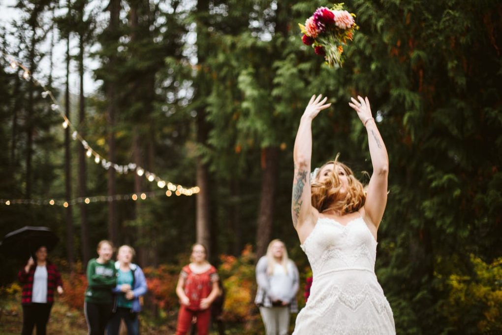 A bride in a white dress throws her bouquet into the air at a snohomish outdoor wedding with several women in the background surrounded by trees and string lights Snohomish wedding photography A bride in a white dress throws her bouquet into the air at a snohomish outdoor wedding with several women in the background surrounded by trees and string lights Snohomish wedding photography