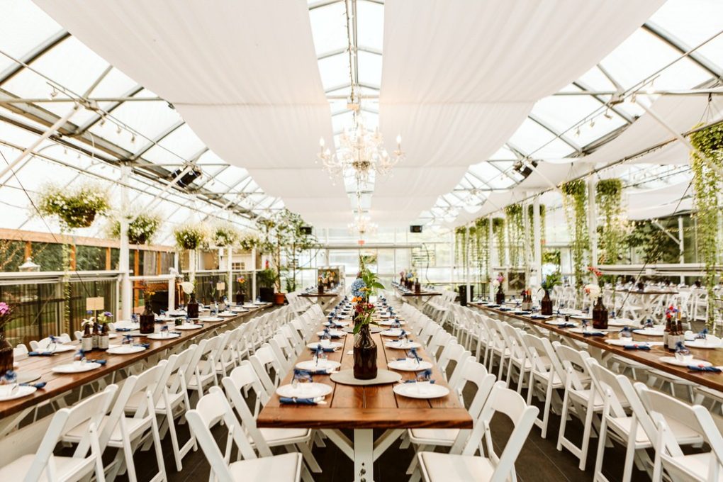 A long banquet table set for an event in a bright snohomish greenhouse-style venue with white chairs wooden tables hanging plants chandeliers and natural light streaming through glass ceilings Snohomish wedding photography A long banquet table set for an event in a bright snohomish greenhouse style venue with white chairs wooden tables hanging plants chandeliers and natural light streaming through glass ceilings Snohomish wedding photography