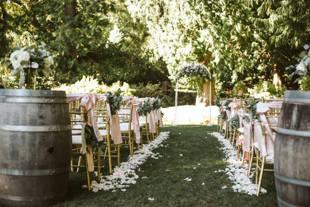 Outdoor wedding ceremony setup in snohomish with gold chairs decorated with pink ribbons flower arrangements and flower petals on the grass aisle leading to a floral arch under trees Snohomish wedding photography Outdoor wedding ceremony setup in snohomish with gold chairs decorated with pink ribbons flower arrangements and flower petals on the grass aisle leading to a floral arch under trees Snohomish wedding photography