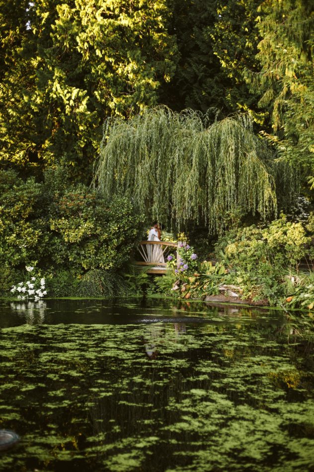 A person sits on a wooden bench beneath a large weeping willow tree in a lush green snohomish garden by a pond covered with floating plants and surrounded by dense foliage and blooming flowers Snohomish wedding photography Washington wedding venue chateau lill