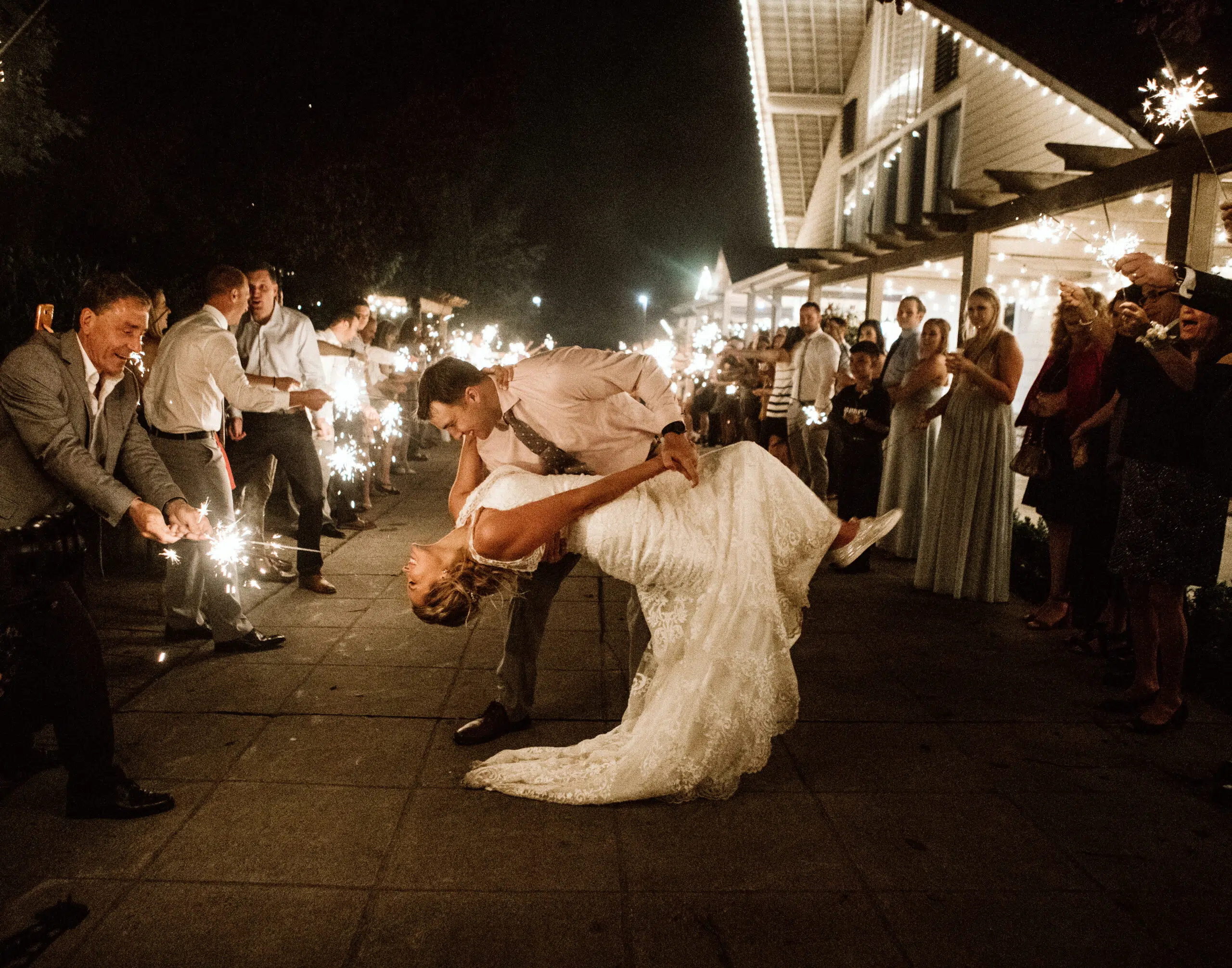 A bride and groom share a dramatic dance dip surrounded by guests holding sparklers at night outside a brightly lit Snohomish venue. The atmosphere is festive and celebratory. Snohomish Wedding Photography