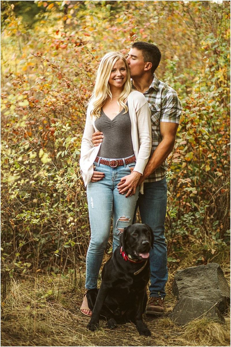 A smiling woman stands in front of a man who kisses her head as they pose outdoors amid vibrant snohomish autumn foliage A black dog wearing a red collar sits at their feet all appearing happy and relaxed Snohomish wedding photography