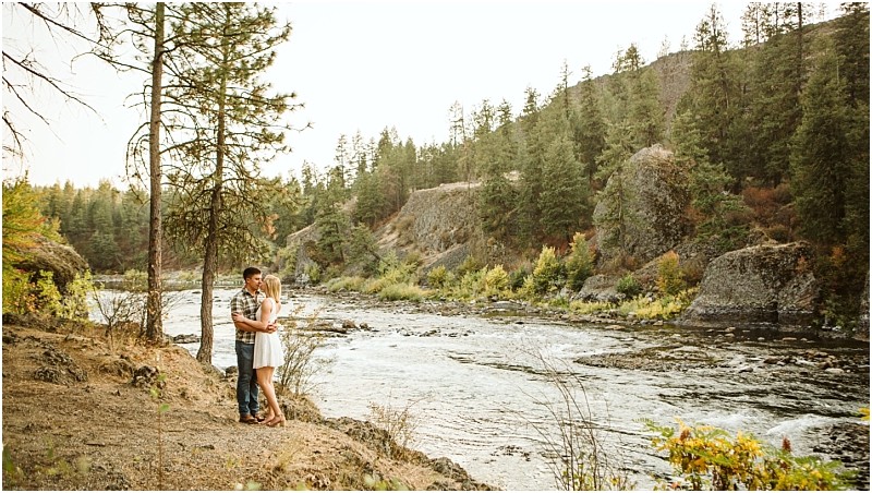 A couple embraces by a tree on a rocky snohomish riverbank surrounded by pine trees and greenery with a flowing river and forested hills in the background Snohomish wedding photography