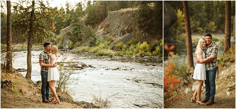 A couple stands embracing by a flowing river in a forested snohomish area The woman wears a white dress and the man a plaid shirt and jeans Trees and rocky terrain surround them as they smile at the camera in warm natural light Snohomish wedding photography