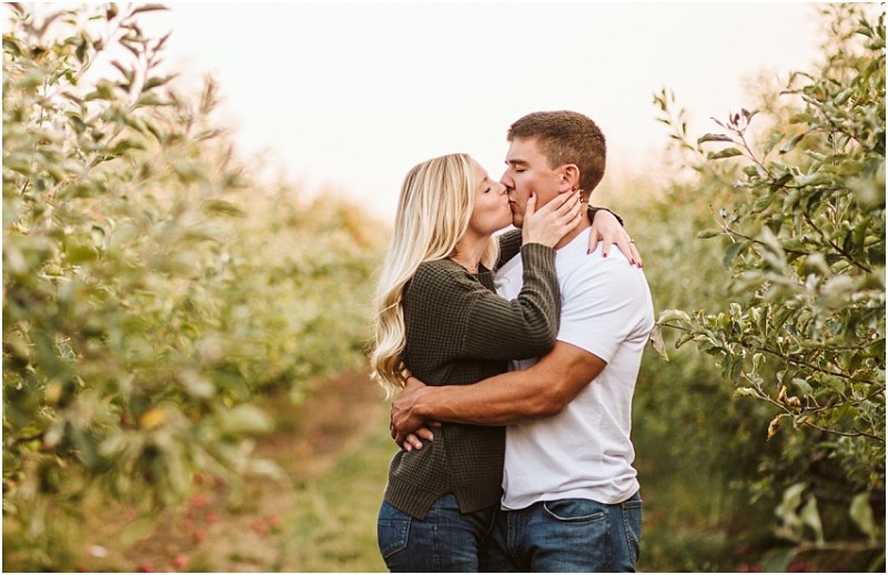 A couple stands in a lush snohomish orchard embracing and kissing The woman with long blonde hair and a dark sweater holds the mans face as he wraps his arms around her Verdant greenery surrounds them Snohomish wedding photography