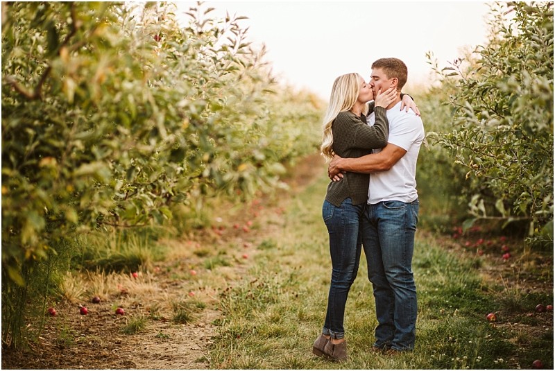 A couple stands in a snohomish apple orchard embracing and kissing They are surrounded by green trees and scattered apples on the ground creating a romantic and natural setting Snohomish wedding photography