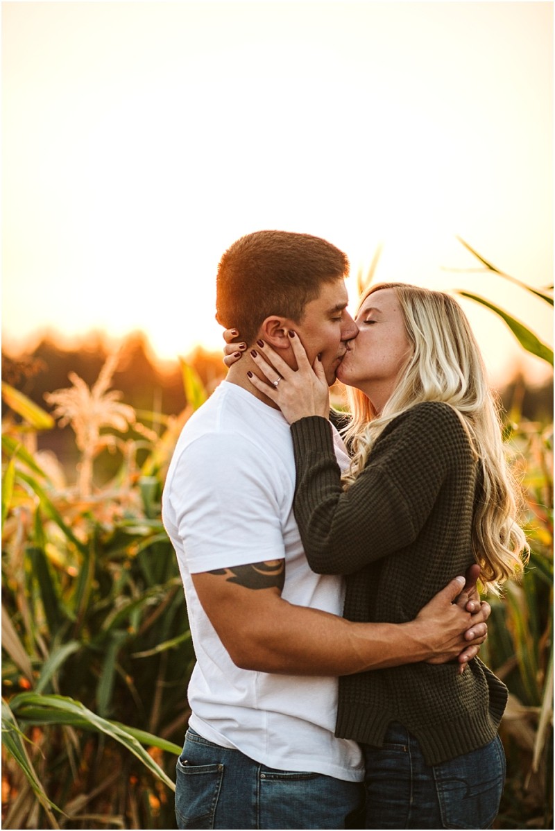A couple stands in a snohomish cornfield at sunset embracing and kissing The man wears a white t shirt and the woman with long blonde hair wears a dark sweater Warm sunlight illuminates the scene Snohomish wedding photography