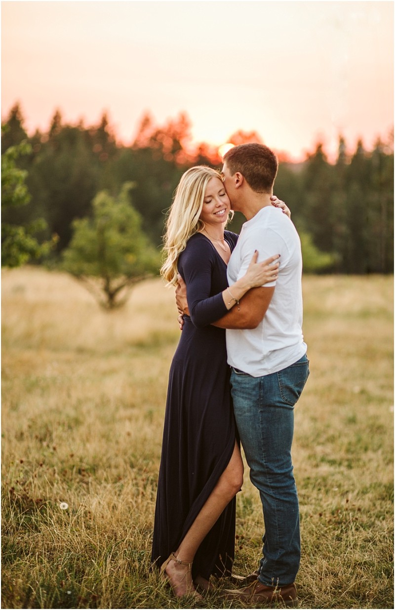A couple stands embracing in a grassy snohomish field at sunset The woman smiling in a long navy dress is held by a man in a white shirt and jeans Trees and a colorful sky paint the background Snohomish wedding photography