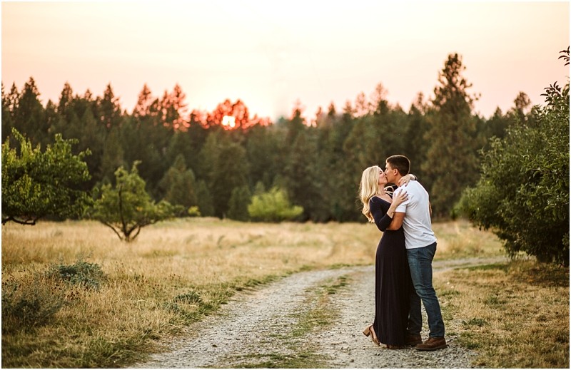A couple embraces and kisses on a dirt path through a grassy snohomish field surrounded by trees with the sun setting behind the forest in the background Snohomish wedding photography