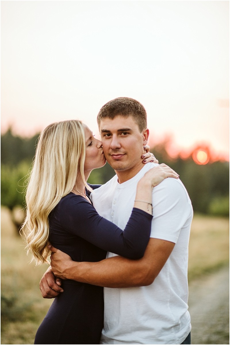 A woman with long blonde hair kisses a man on the cheek while hugging him outdoors at sunset in snohomish The man in a white shirt looks at the camera as the woman holds him closely against a blurred field and trees Snohomish wedding photography