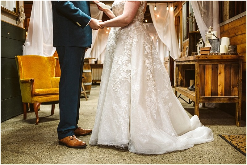 A bride and groom stand facing each other holding hands in a warmly lit snohomish venue The bride wears a white lace gown the groom a blue suit They are near a yellow armchair and wooden furniture creating an intimate setting Snohomish wedding photography