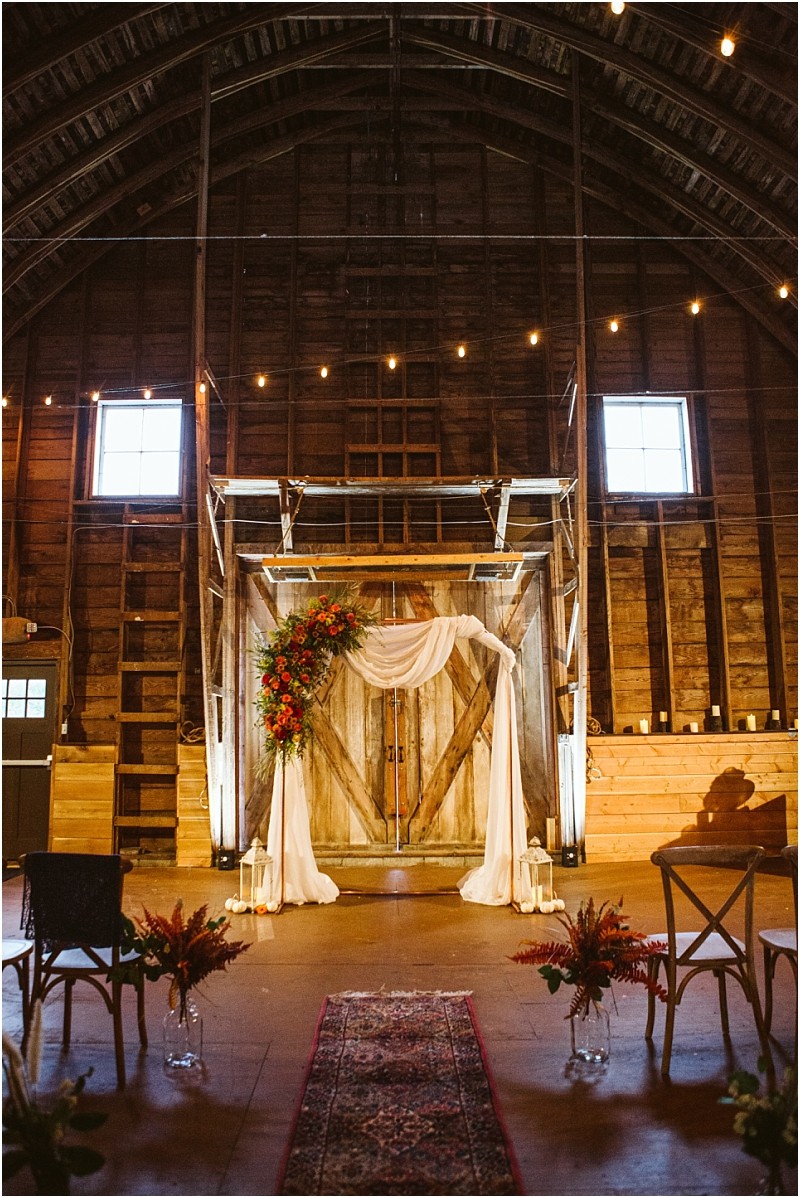 A rustic snohomish barn interior set up for a wedding ceremony with draped white fabric and colorful floral arrangements on wooden doors a patterned rug and string lights hanging above Snohomish wedding photography
