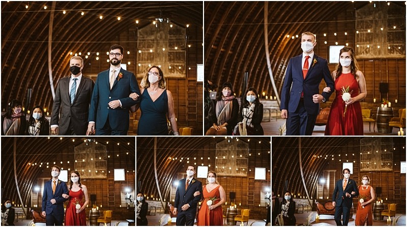 A wedding party walks down the aisle in a rustic snohomish barn venue All participants and guests wear face masks women in red dresses and men in suits beneath warm string lights that hang overhead Snohomish wedding photography