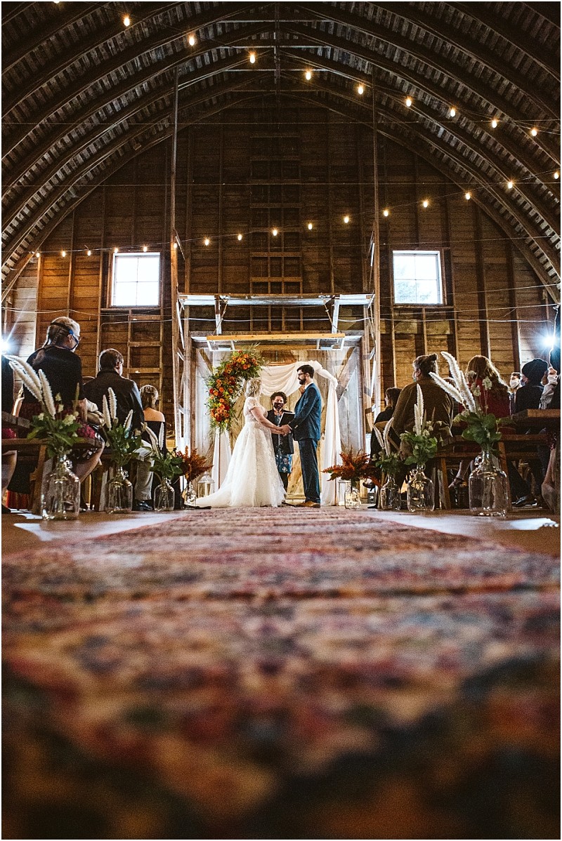 A bride and groom stand together at the altar in a rustic snohomish barn wedding venue surrounded by guests on either side The barn is beautifully decorated with string lights and floral arrangements Snohomish wedding photography