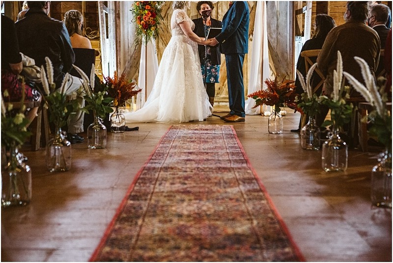 A couple stands holding hands at the altar during their snohomish wedding ceremony with a celebrant in the background Guests are seated on either side of an aisle decorated with flowers in glass jars Snohomish wedding photography