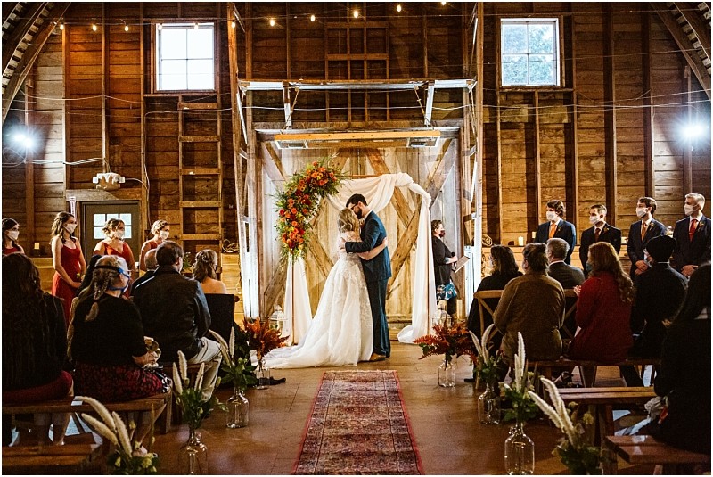 A bride and groom kiss at the altar in a rustic snohomish barn wedding ceremony surrounded by their wedding party and guests seated on benches with warm lighting and floral decorations Snohomish wedding photography
