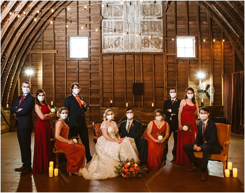 A wedding party poses in a rustic snohomish barn all wearing face masks The bride and groom sit surrounded by bridesmaids in red dresses and groomsmen in dark suits String lights and candles add a warm cozy atmosphere Snohomish wedding photography