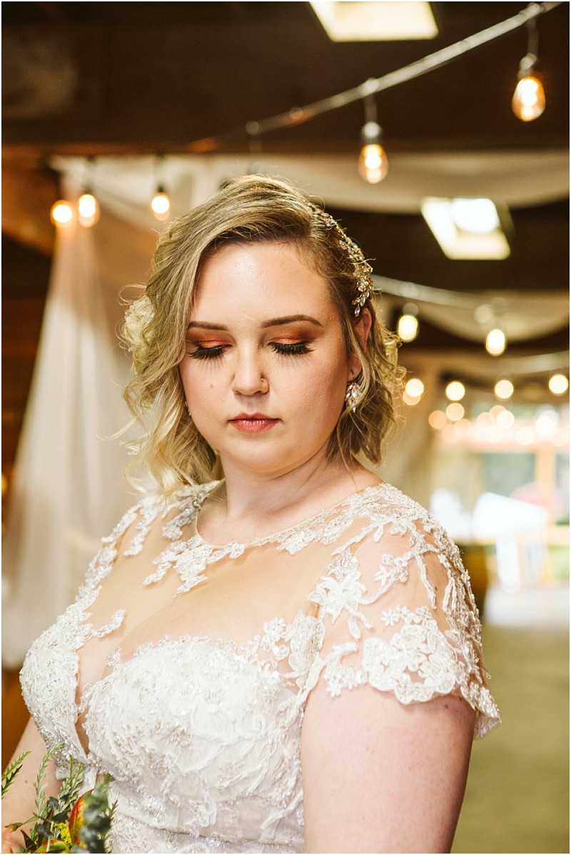 A bride with short blonde hair and soft makeup stands indoors in a lace wedding dress looking down with a calm expression Warm string lights and draped fabric create a dreamy snohomish backdrop Snohomish wedding photography