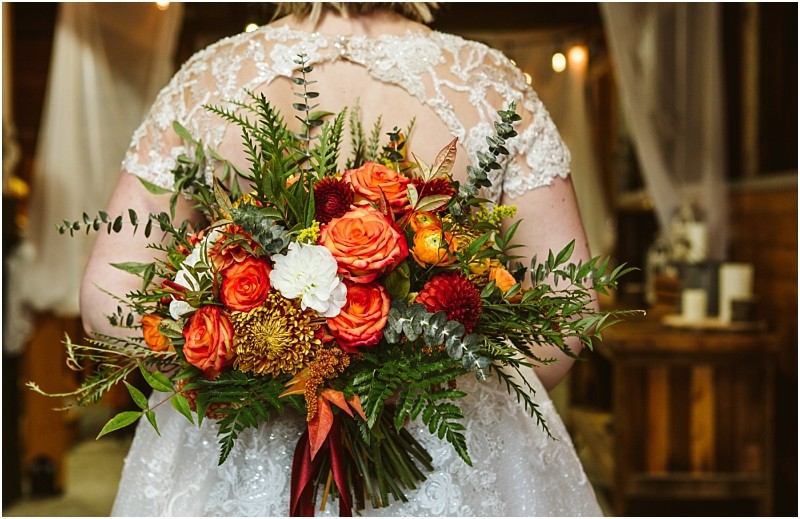 A bride in a lace dress holds a large bouquet of orange red and white flowers with greenery viewed from behind in a warmly lit rustic snohomish setting Snohomish wedding photography