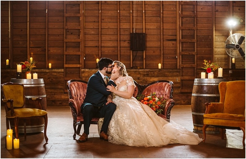 A bride and groom share a kiss while seated on a vintage sofa in a rustic warmly lit snohomish barn decorated with candles flowers and wooden barrels The bride wears a white gown and the groom wears a dark suit Snohomish wedding photography