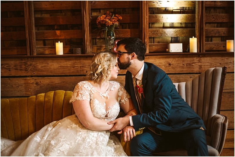 A bride and groom sit closely on a sofa holding hands The groom kisses the brides forehead Indoors in snohomish candles and a vase of flowers rest on a rustic wooden background behind them Snohomish wedding photography