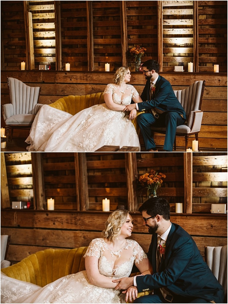 A bride in a lace wedding gown and a groom in a suit sit together on a couch in a rustic wood paneled snohomish room smiling and holding hands Candles and flowers decorate the cozy background Snohomish wedding photography