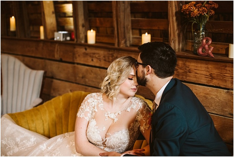 A bride in a lace wedding dress sits on a yellow couch in snohomish eyes closed as a groom in a dark suit gently kisses her temple Candlelight and rustic wooden decor create a warm intimate atmosphere Snohomish wedding photography