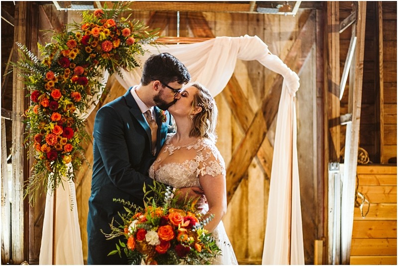 A bride and groom kiss under a wooden arch decorated with white fabric and vibrant orange flowers standing in a rustic snohomish barn setting The bride holds a bouquet of matching flowers Snohomish wedding photography