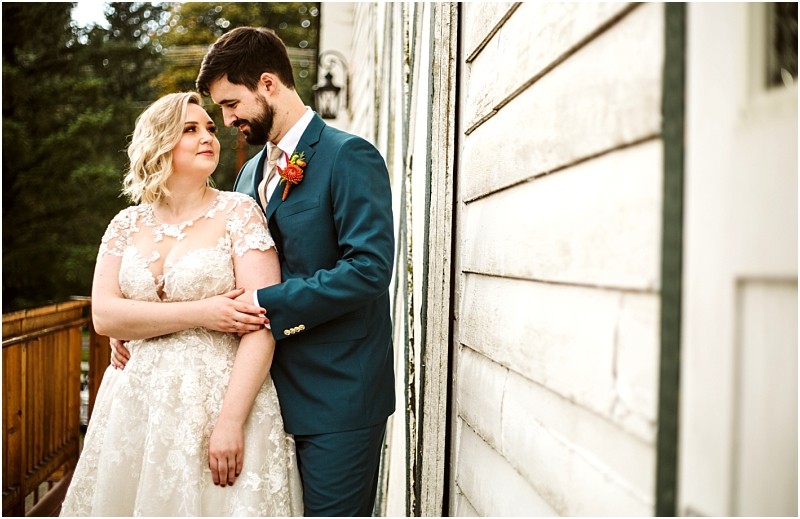 A bride in a lace wedding dress and a groom in a blue suit stand closely together on a wooden deck in snohomish smiling at each other with a rustic white building beside them Snohomish wedding photography
