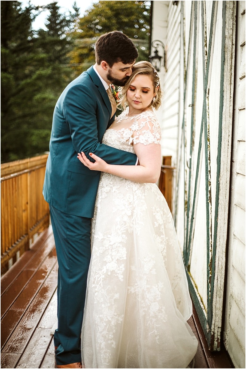 A groom in a teal suit gently kisses his brides forehead as they embrace on a wooden deck beside a white building with green trim in snohomish with trees visible in the background Snohomish wedding photography