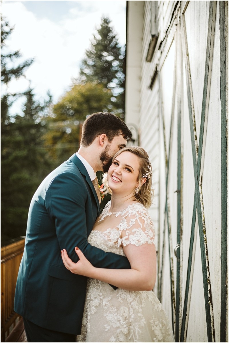 A groom in a teal suit kisses his smiling bride who wears a lace wedding dress as they embrace beside a white wooden building with green trim in snohomish Trees and blue sky are visible in the background Snohomish wedding photography