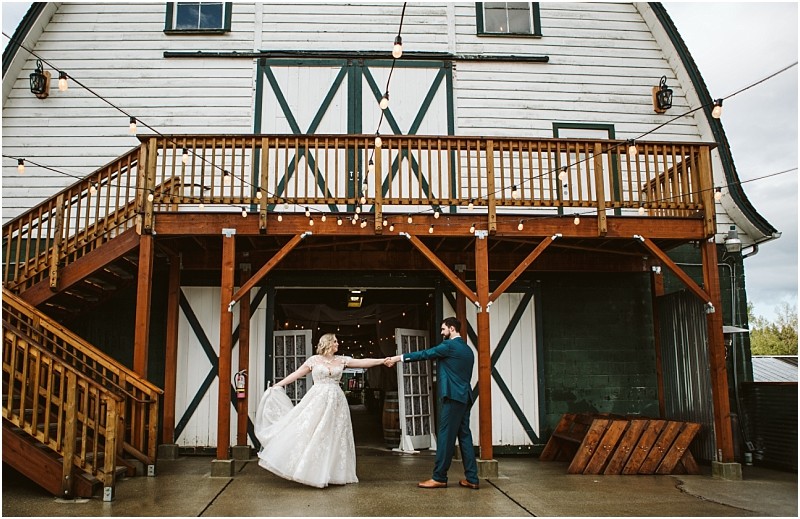 A bride and groom dance outside a rustic snohomish barn with wooden railings and string lights the bride in a white gown and the groom in a blue suit holding hands and smiling Snohomish wedding photography