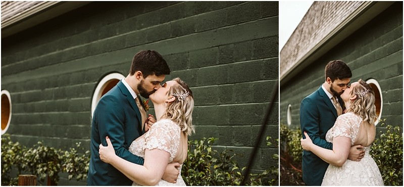 A bride and groom embrace and share a kiss outside standing in front of a green brick wall with round windows and bushes in snohomish The groom wears a dark teal suit the bride wears a white lace dress Snohomish wedding photography