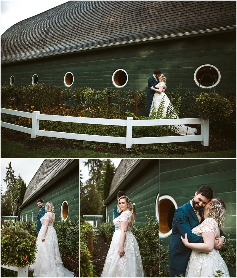 A bride and groom pose together outside a green barn with round windows and a white fence in snohomish surrounded by gardens The bride wears a lace gown and the groom is in a blue suit They look happy and affectionate Snohomish wedding photography