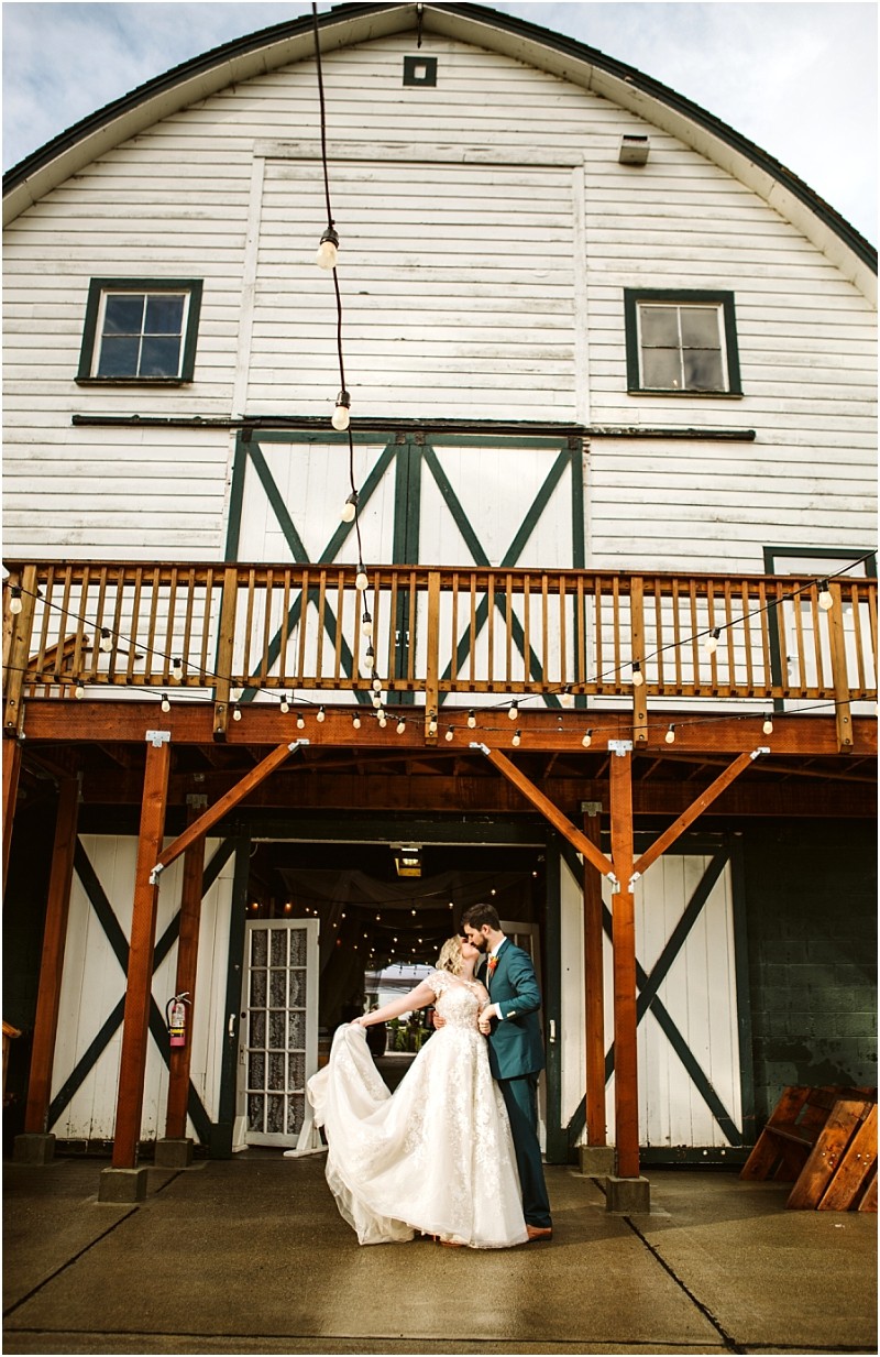 A bride in a white gown and a groom in a blue suit share a kiss and dance outside a rustic snohomish white barn with green trim and wooden balcony on a cloudy day String lights hang above them Snohomish wedding photography